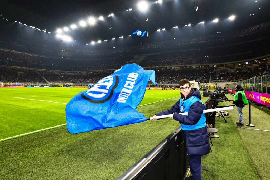 Giovane sbandieratore Inter Club a San Siro con bandiera neroazzurra e stemma Inter, in campo durante una partita di Serie A di calcio, stadio Giuseppe Meazza pieno di tifosi.